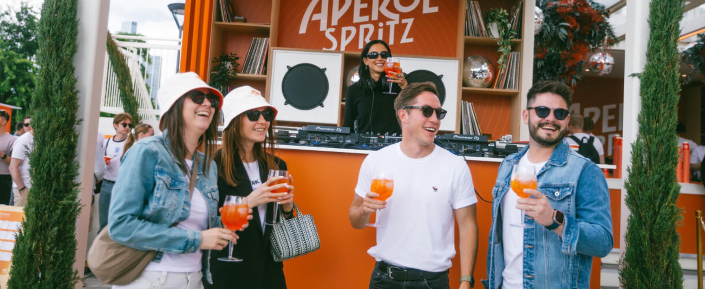 4 people stand holding aperol Spritz with a DJ playing behind them