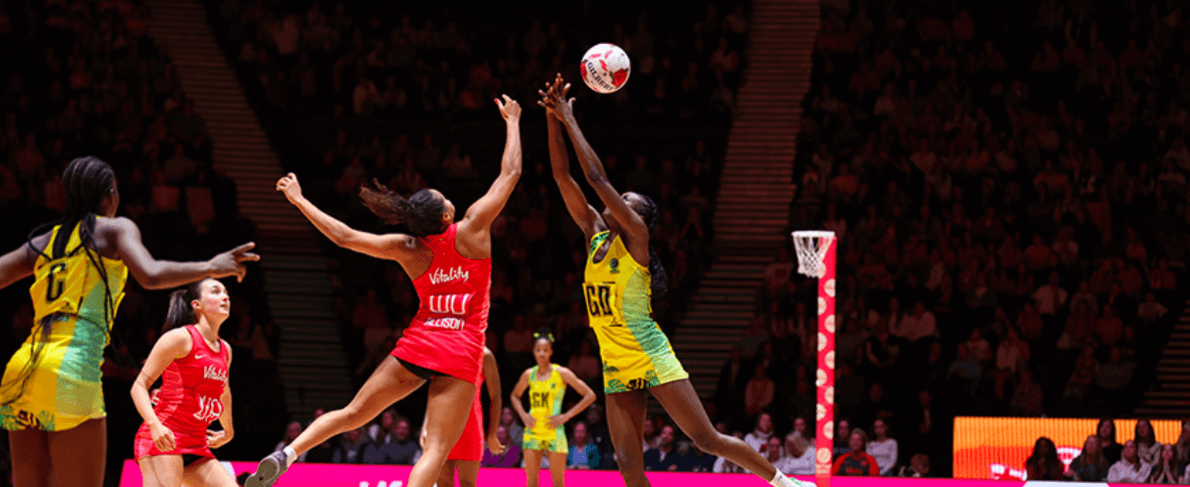 Two competing netball teams in yellow and pink jerseys play on the court.