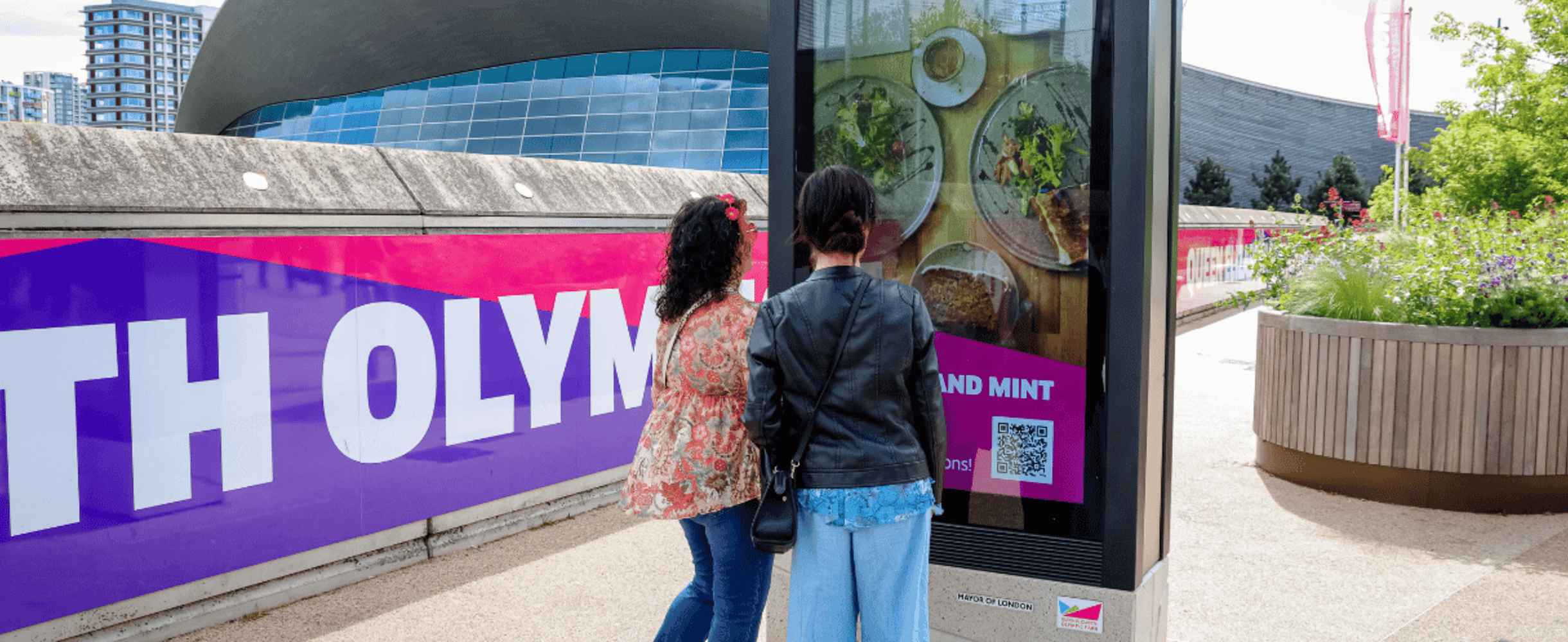 Two women looking at a digital screen advertisement on Queen Elizabeth Olympic Park