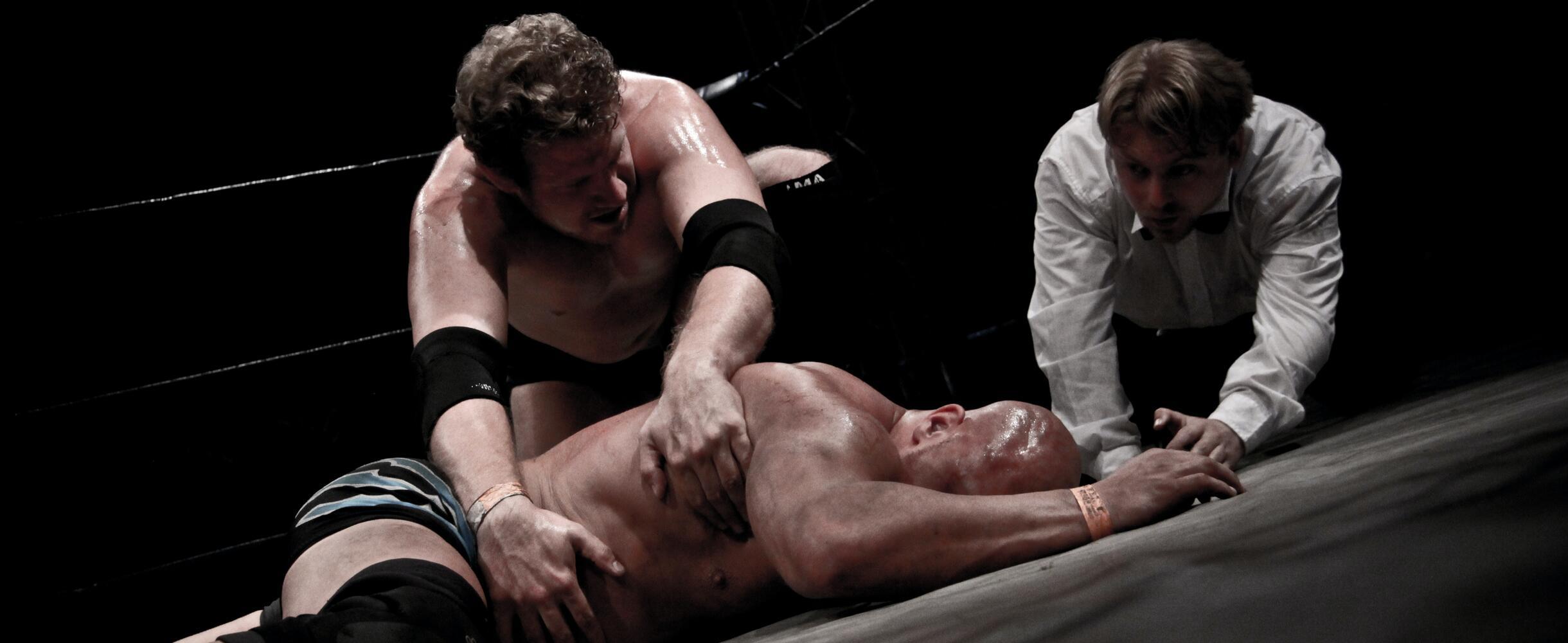 2 men wrestling, 1 lying on ground with referee in background