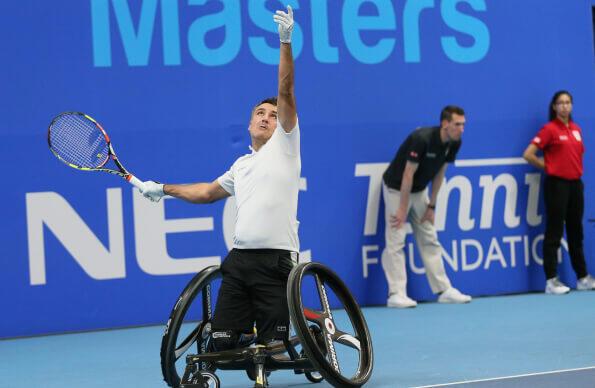 A man in a wheelchair serves during a tennis game