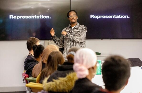 A group of youths listening to an adult presenting a seminar with "representation" written on the board