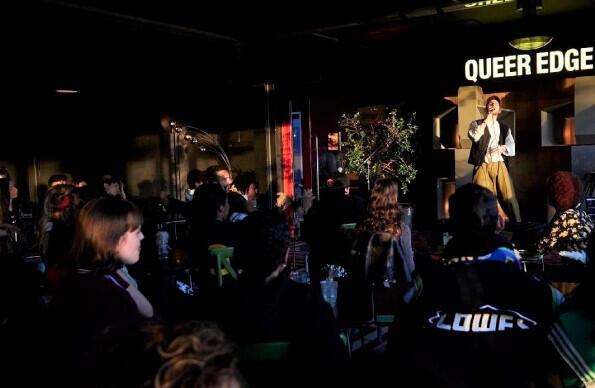 Queer Edge night with person on stage in front of crowd Queer Edge night with person on stage in front of crowd