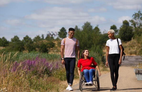 3 people, one in a wheelchair on a path with flowers and Olympic rings in the background