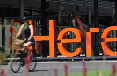 A person cycling past an orange HERE sign on the Here East Campus