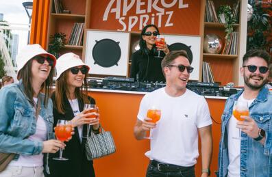 4 people stand holding aperol Spritz with a DJ playing behind them
