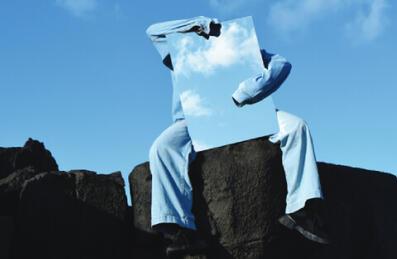 An artist in a blue suit sits on a rock with blue sky behind