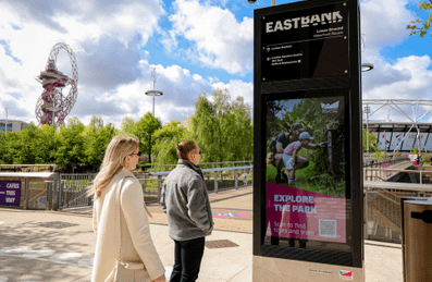 A man and a woman looking at a digital screen advertisement on Queen Elizabeth Olympic Park