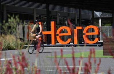 A woman cycling past an orange HERE sign on the Here East campus